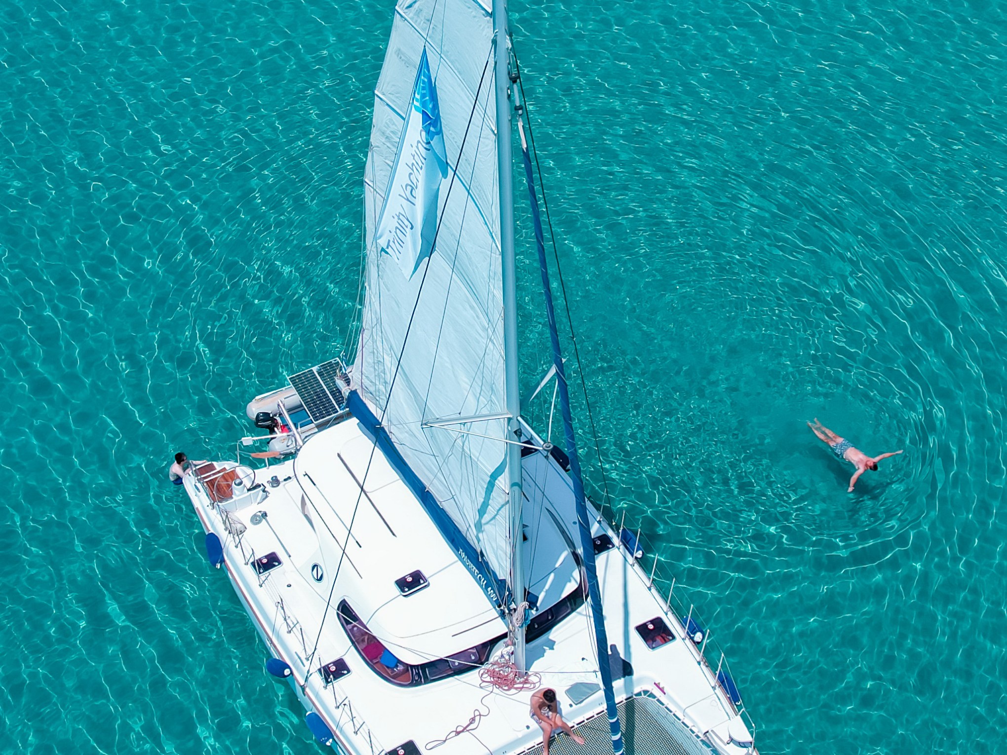 Aerial view of a sailboat with people on clear turquoise water, person swimming nearby.