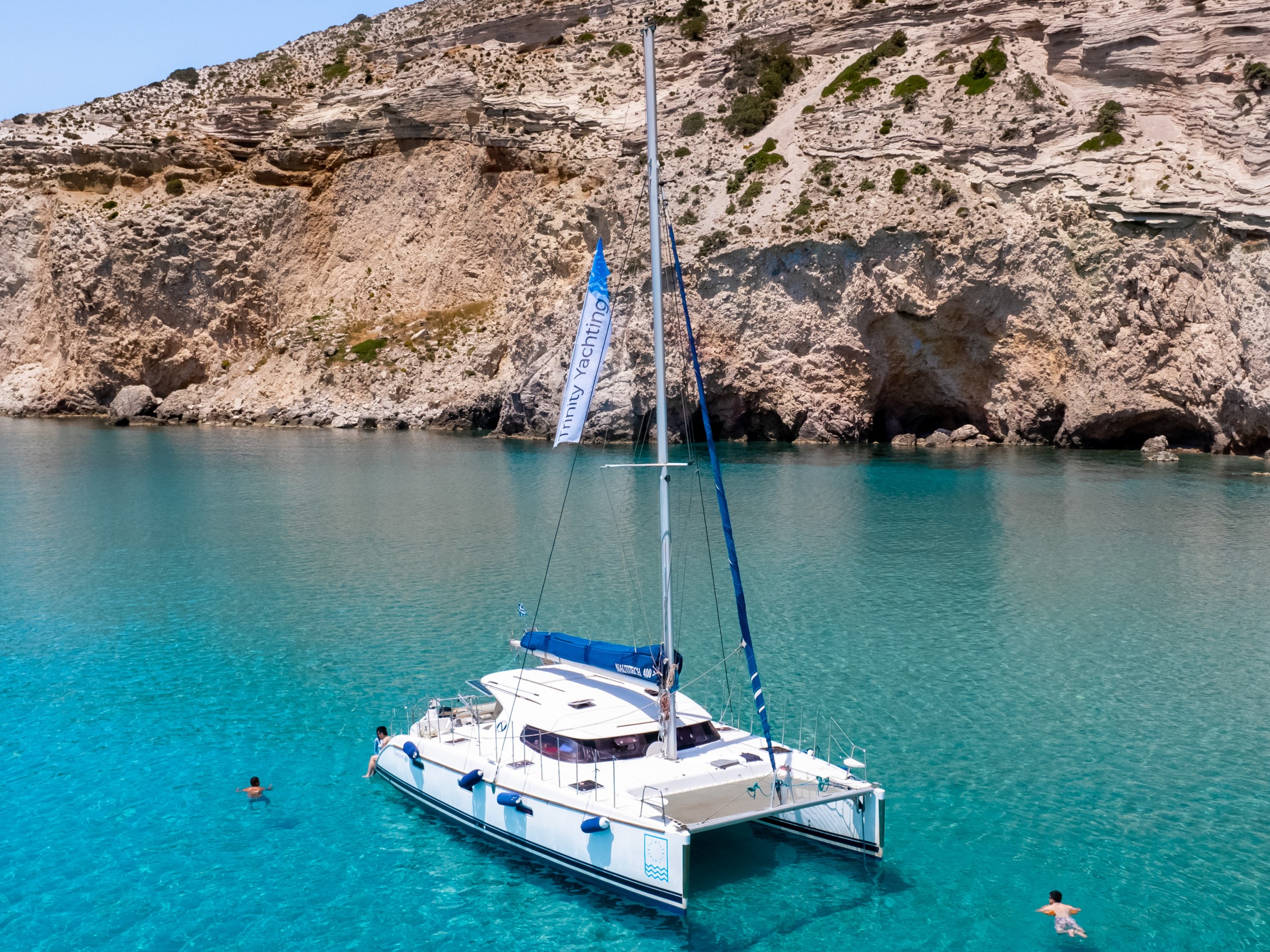 Sailing catamaran in turquoise bay near rocky cliff, swimmers nearby.