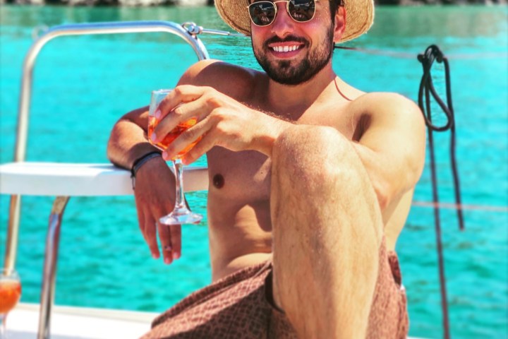 Man in hat and sunglasses relaxes on a boat with a drink, blue water and rocky background.