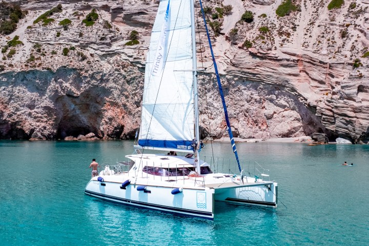 Sailboat in clear turquoise water near rocky cliffs, sunny day, person on boat's deck.