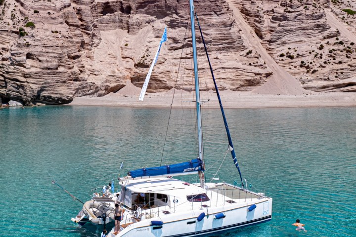 Sailboat on turquoise water near a rocky cliff, with people swimming nearby.