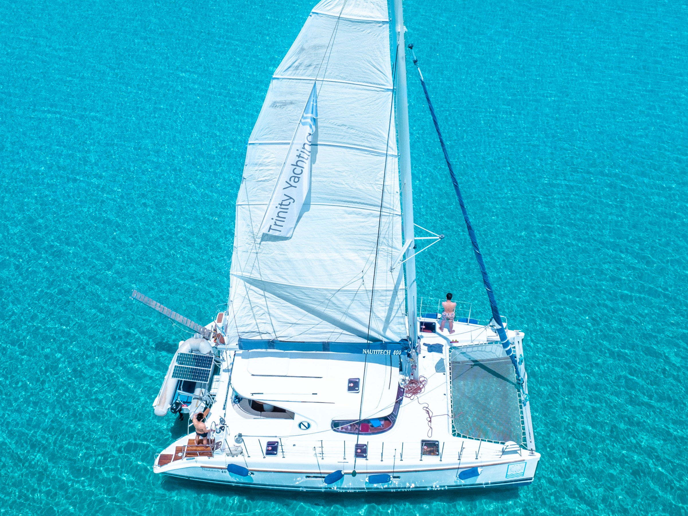 Sailboat with a white sail on clear turquoise water viewed from above.