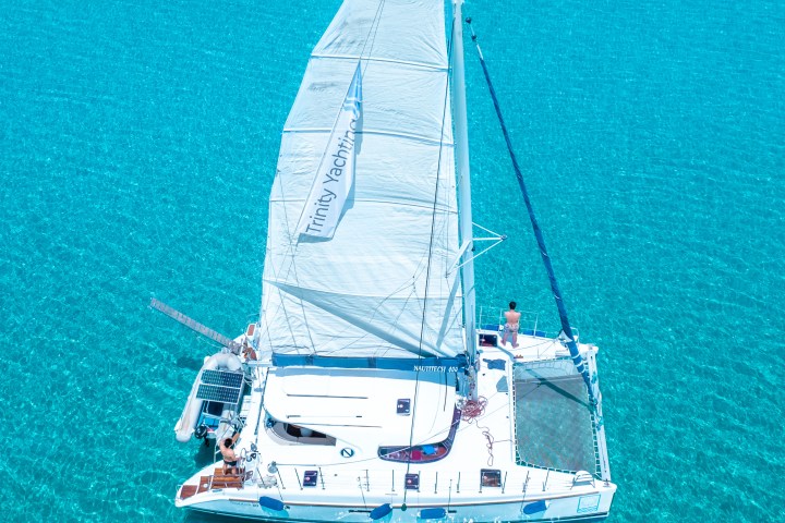 Sailboat with a white sail on clear turquoise water viewed from above.
