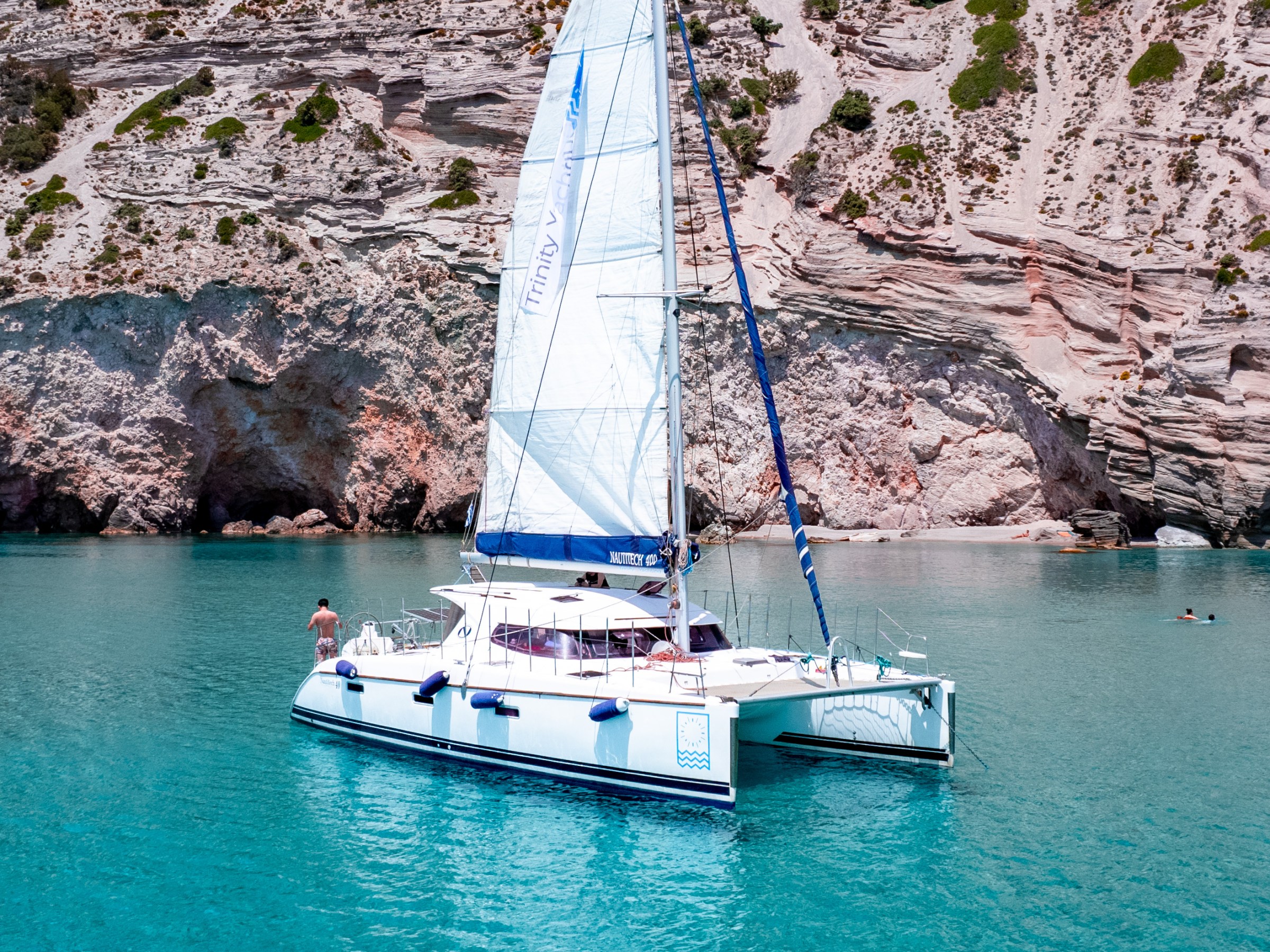 White catamaran with sails in clear blue water near rocky cliffs.