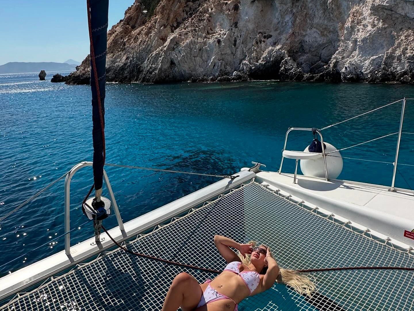 Person in bikini relaxing on a yacht net with rocky coastline in the background.