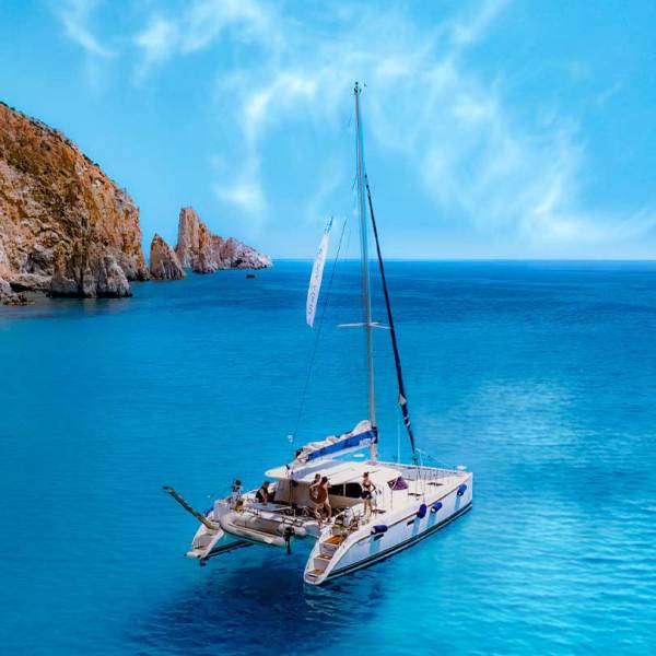 Catamaran sailing near rocky cliffs on calm turquoise water under a blue sky.