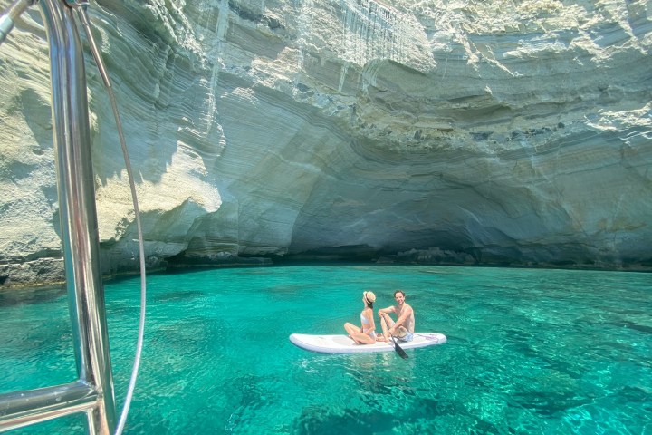 Two people sit on a paddleboard in clear water inside a rocky coastal cave.