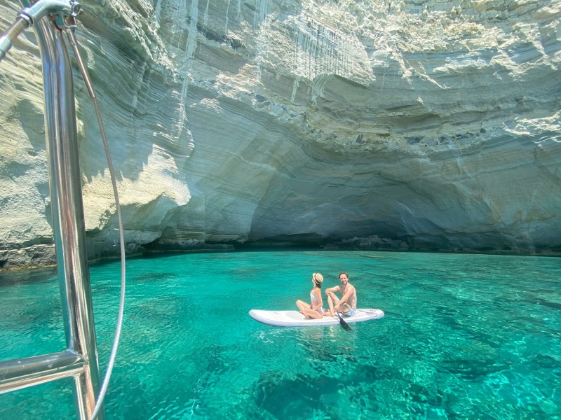 Two people sit on a paddleboard in clear water inside a rocky coastal cave.