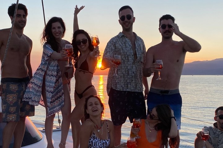 Group of people on a boat at sunset holding drinks, with mountains in the background.