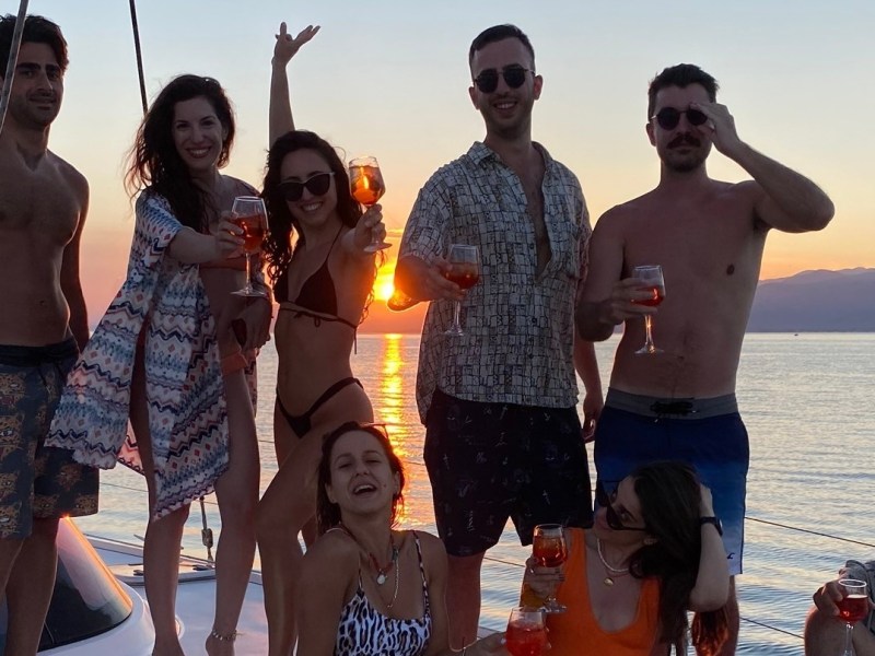 Group of people on a boat at sunset holding drinks, with mountains in the background.