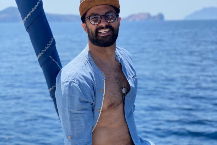 Man in floral shorts and cap smiles on a boat with ocean and islands in background.