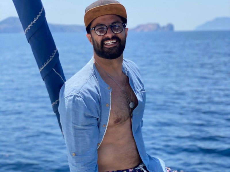 Man in floral shorts and cap smiles on a boat with ocean and islands in background.
