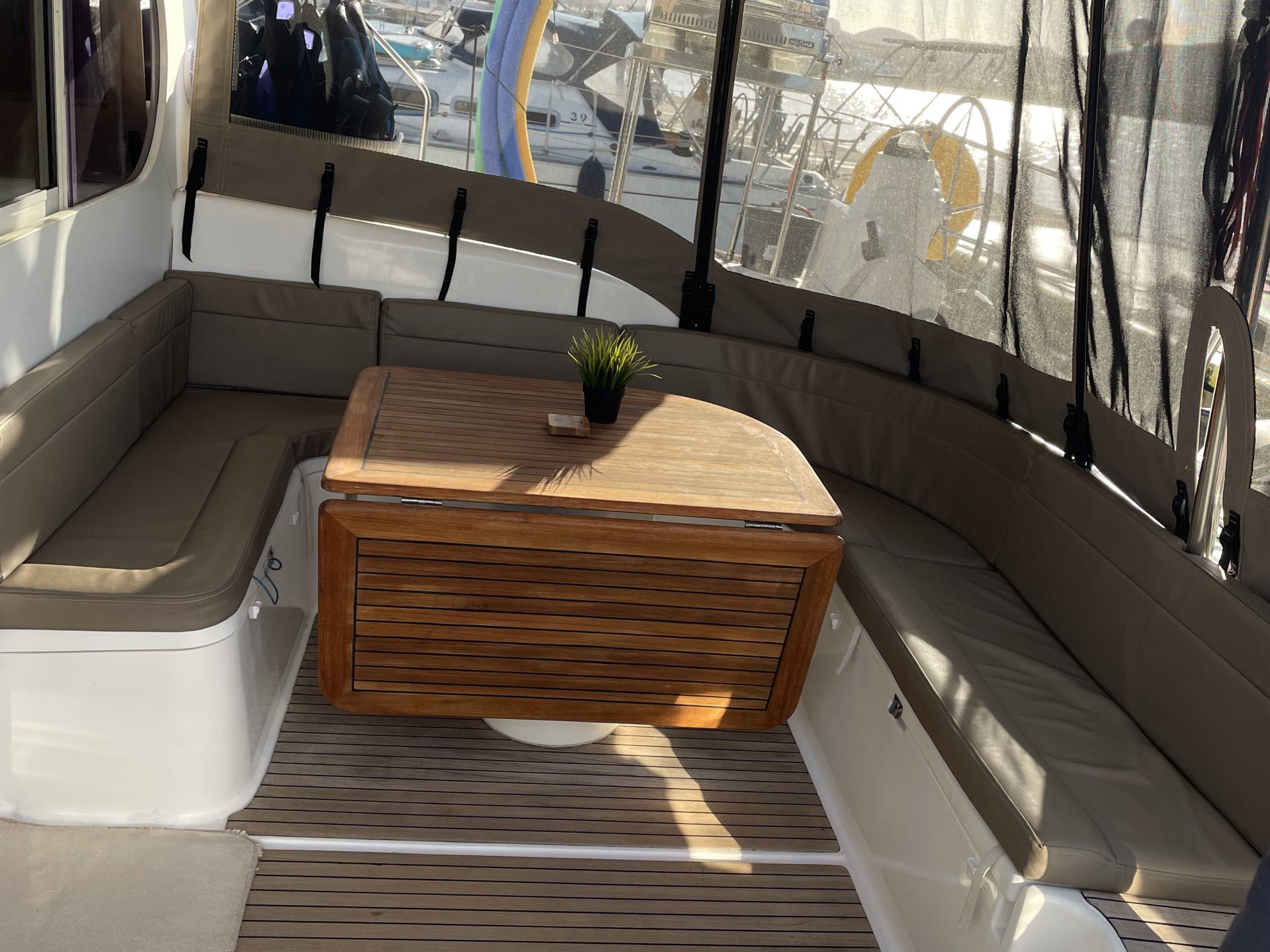 Boat interior with cushioned seating and wooden table, marina visible through windows.