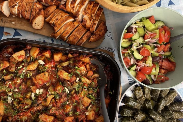 Table with grilled chicken, pasta salad, vegetables, sliced bread, and grape leaves.
