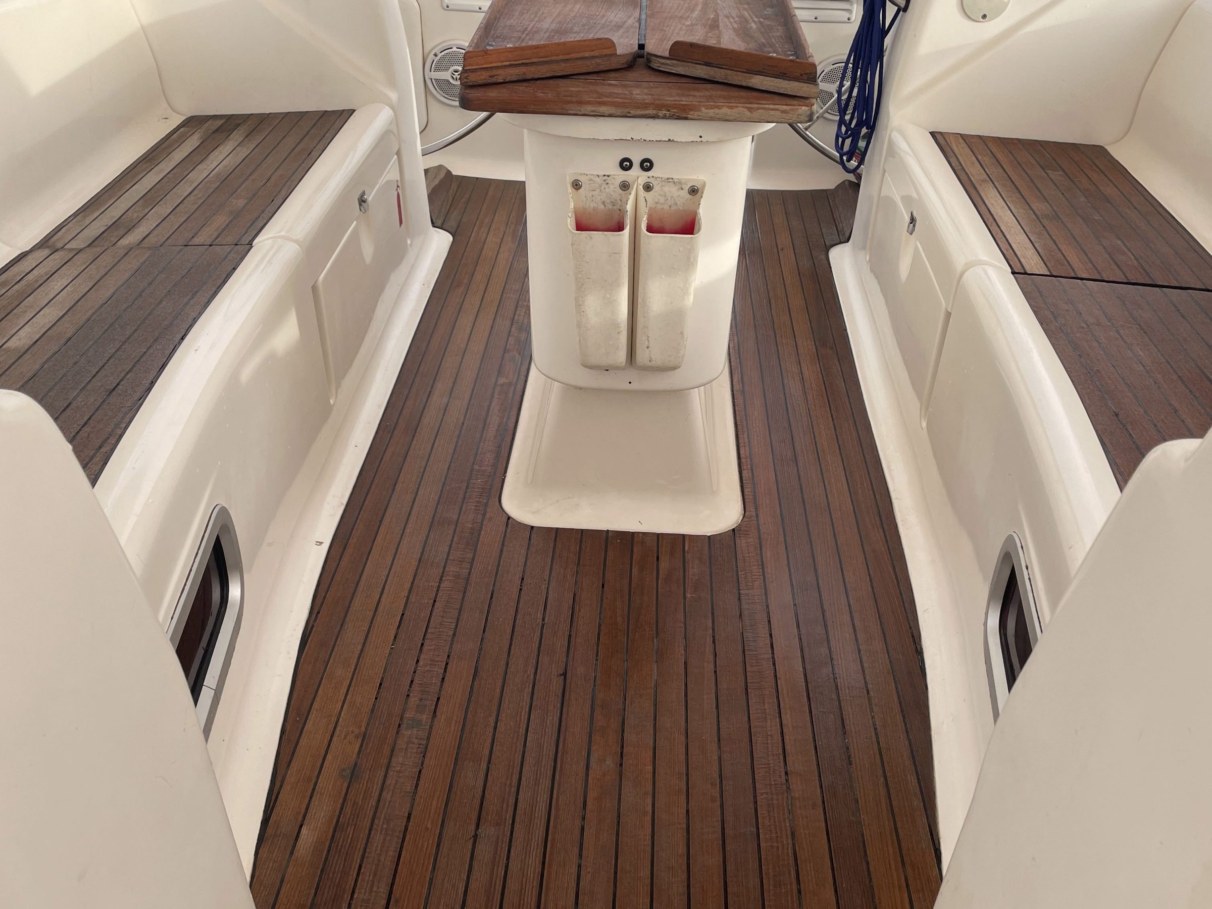 Sailboat cockpit with wooden floor, folding table, and steering wheels on a calm sea backdrop.