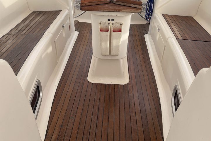 Sailboat cockpit with wooden floor, folding table, and steering wheels on a calm sea backdrop.
