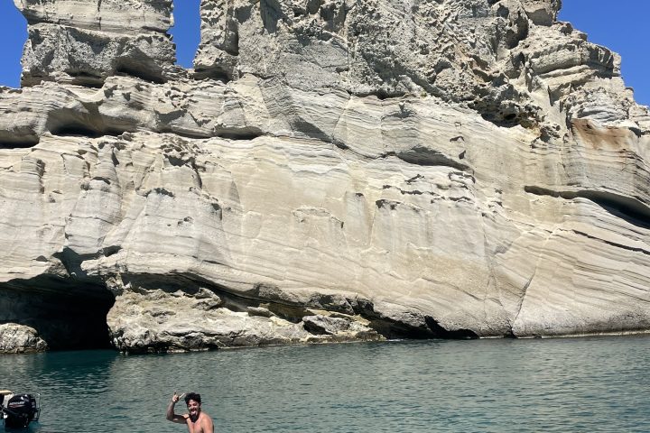 Two people paddleboarding near large rock formations on clear, blue water.