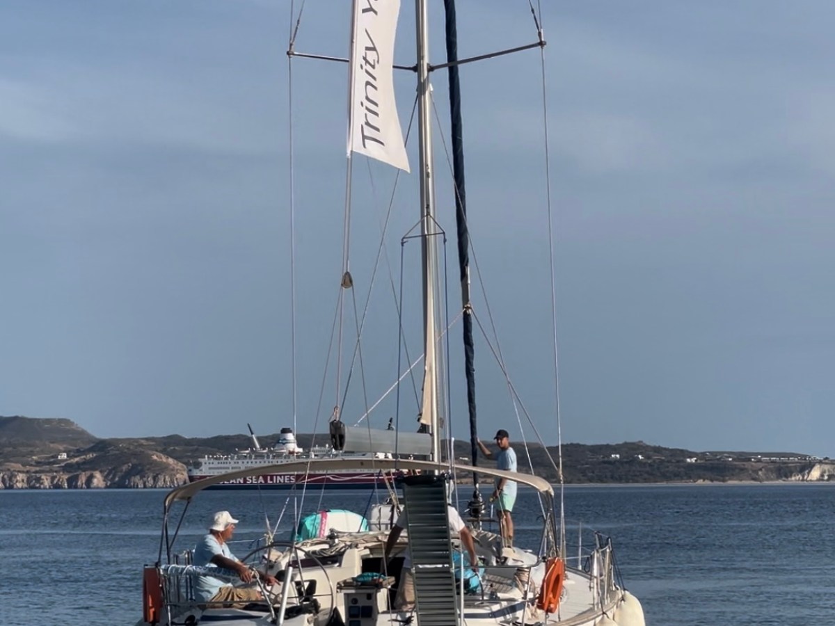 Sailboat with 'Trinity Yachting' banner on calm water, distant hills in background.