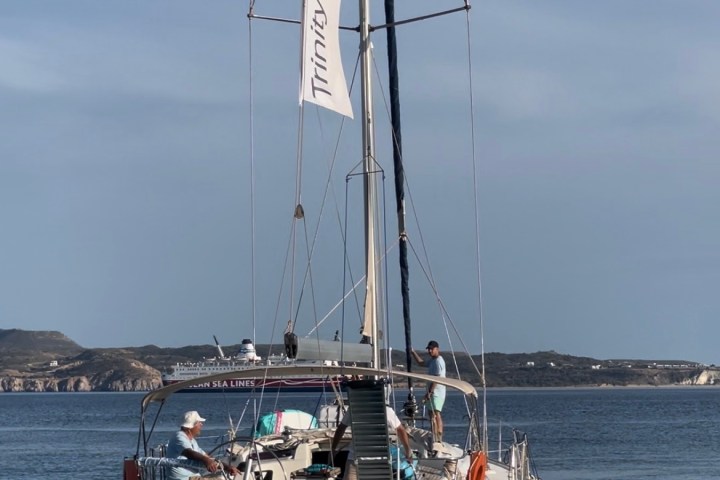 Sailboat with 'Trinity Yachting' banner on calm water, distant hills in background.