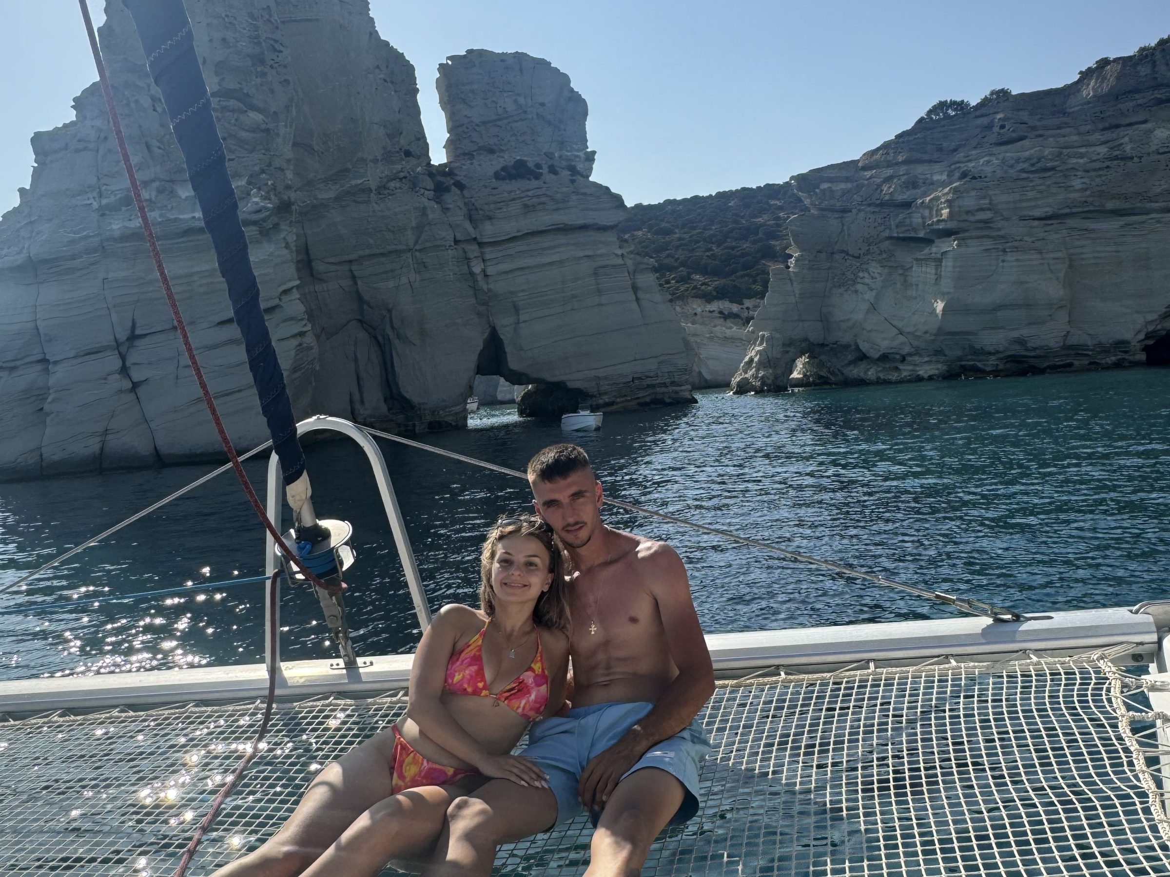 Couple relaxing on a boat's netting with rocky cliffs in the background.