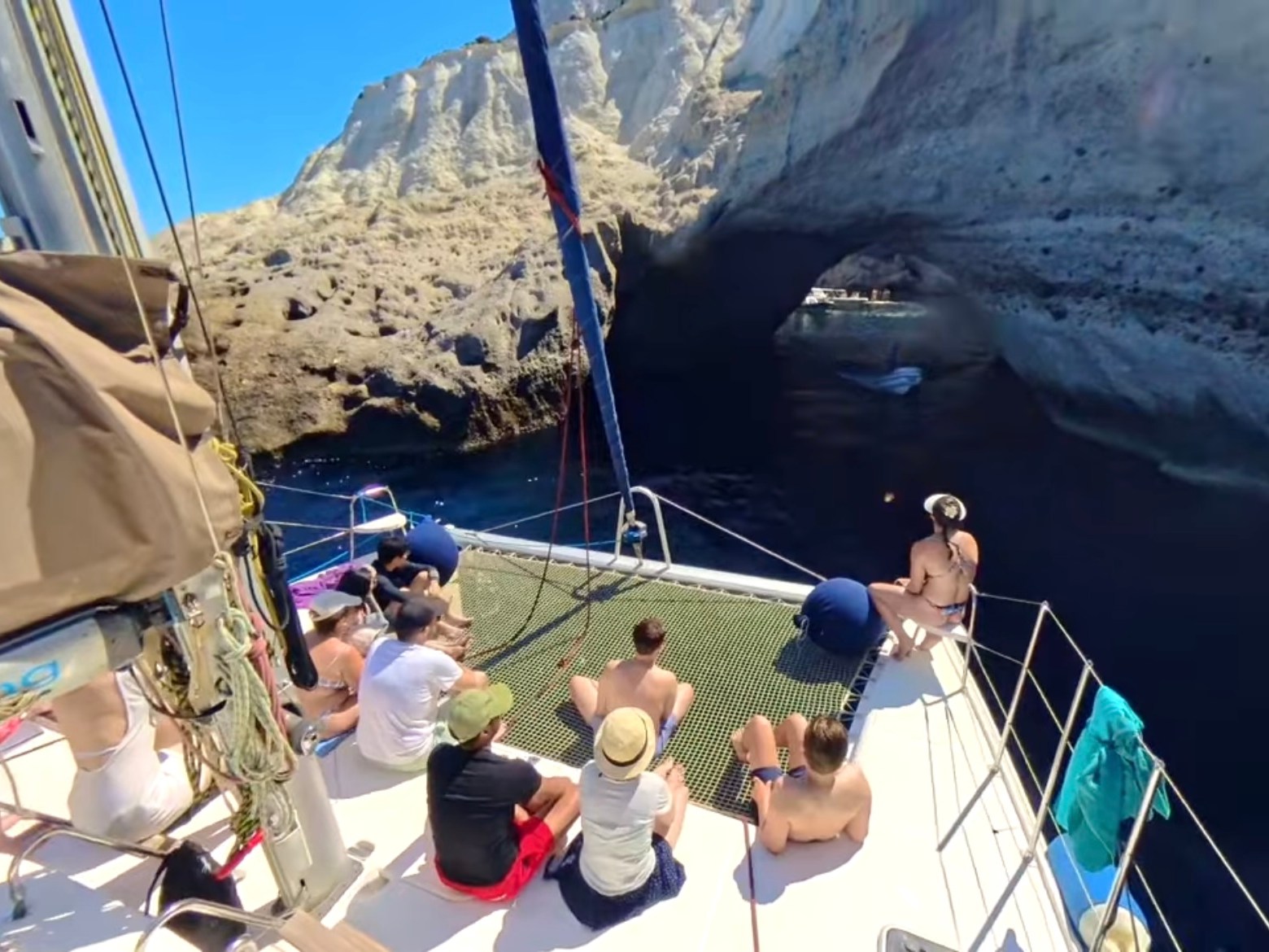 People on a sailboat approach a rocky sea arch in sunny weather.