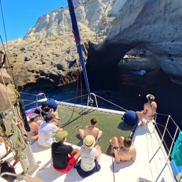People on a sailboat approach a rocky sea arch in sunny weather.