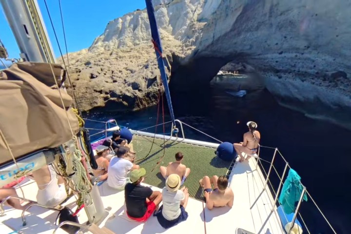 People on a sailboat approach a rocky sea arch in sunny weather.