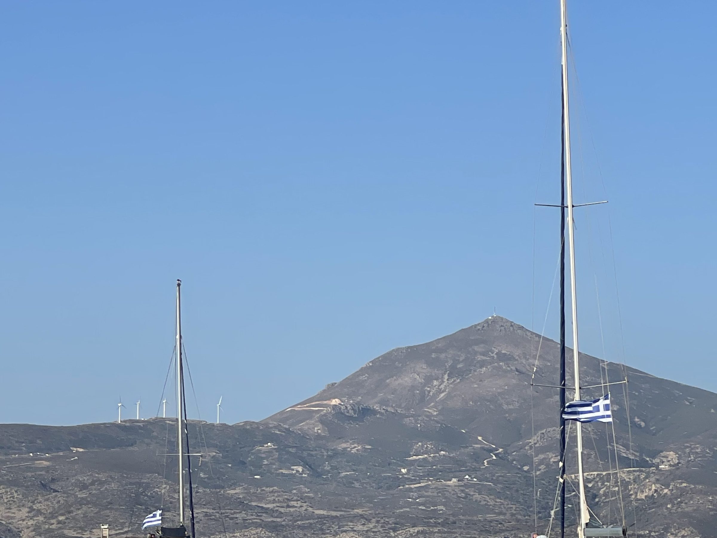 Two sailboats with Greek flags on the sea, mountain in the background under a clear blue sky.