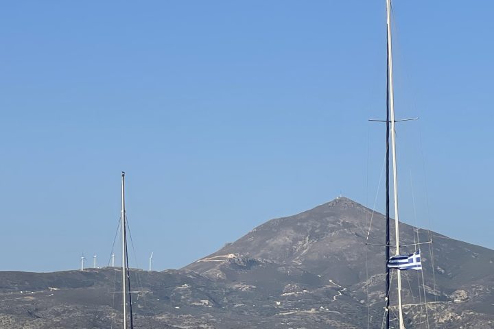 Two sailboats with Greek flags on the sea, mountain in the background under a clear blue sky.