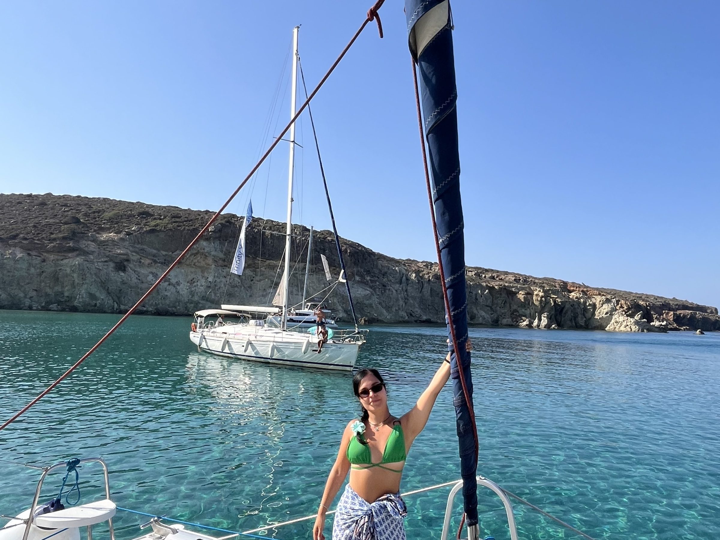 Woman on a boat in clear water with a distant sailboat and rocky shoreline.