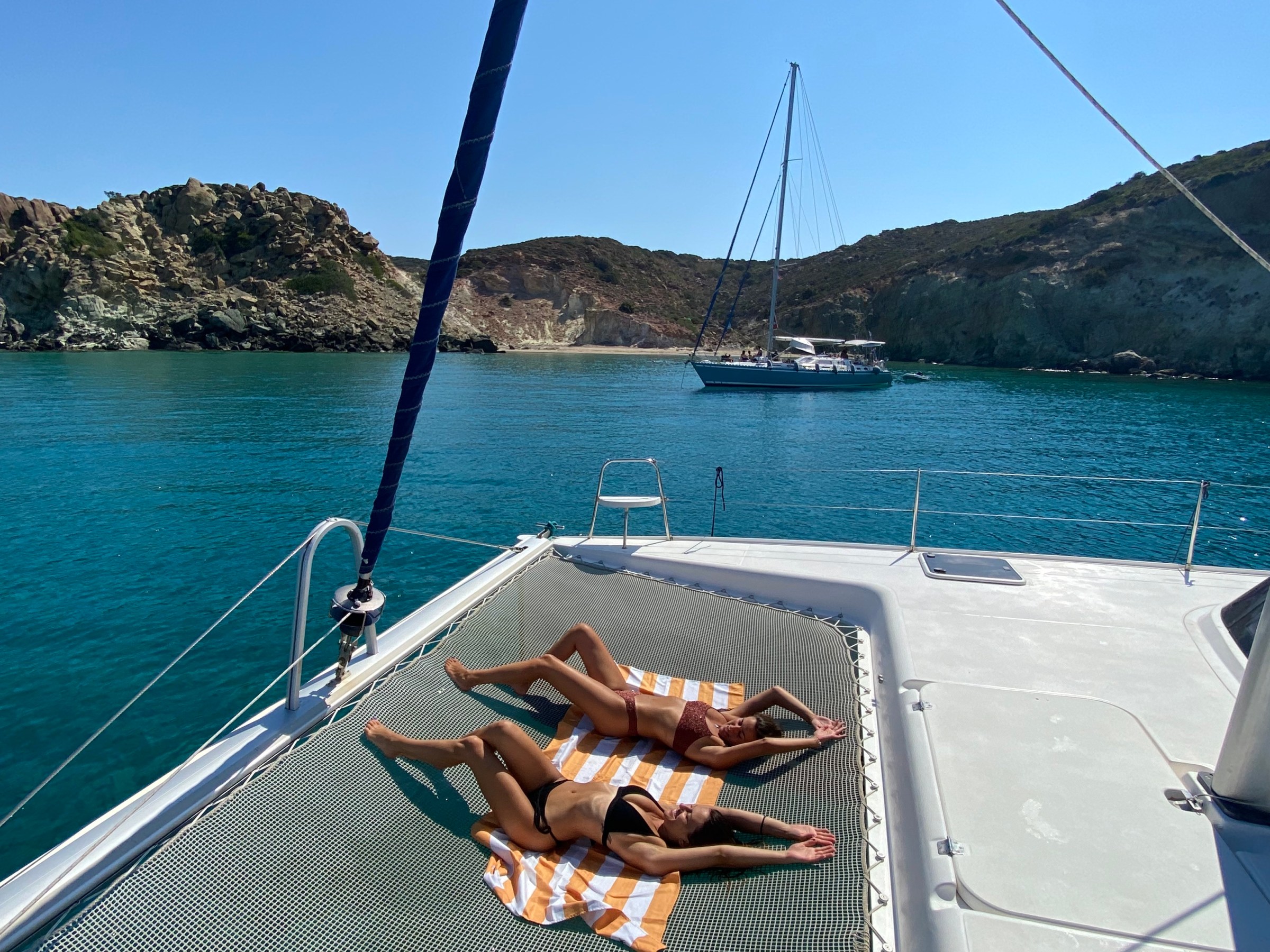 Two people sunbathing on a yacht deck with a sailboat and rocky shore in the background.