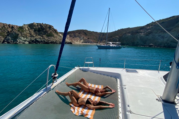 Two people sunbathing on a yacht deck with a sailboat and rocky shore in the background.