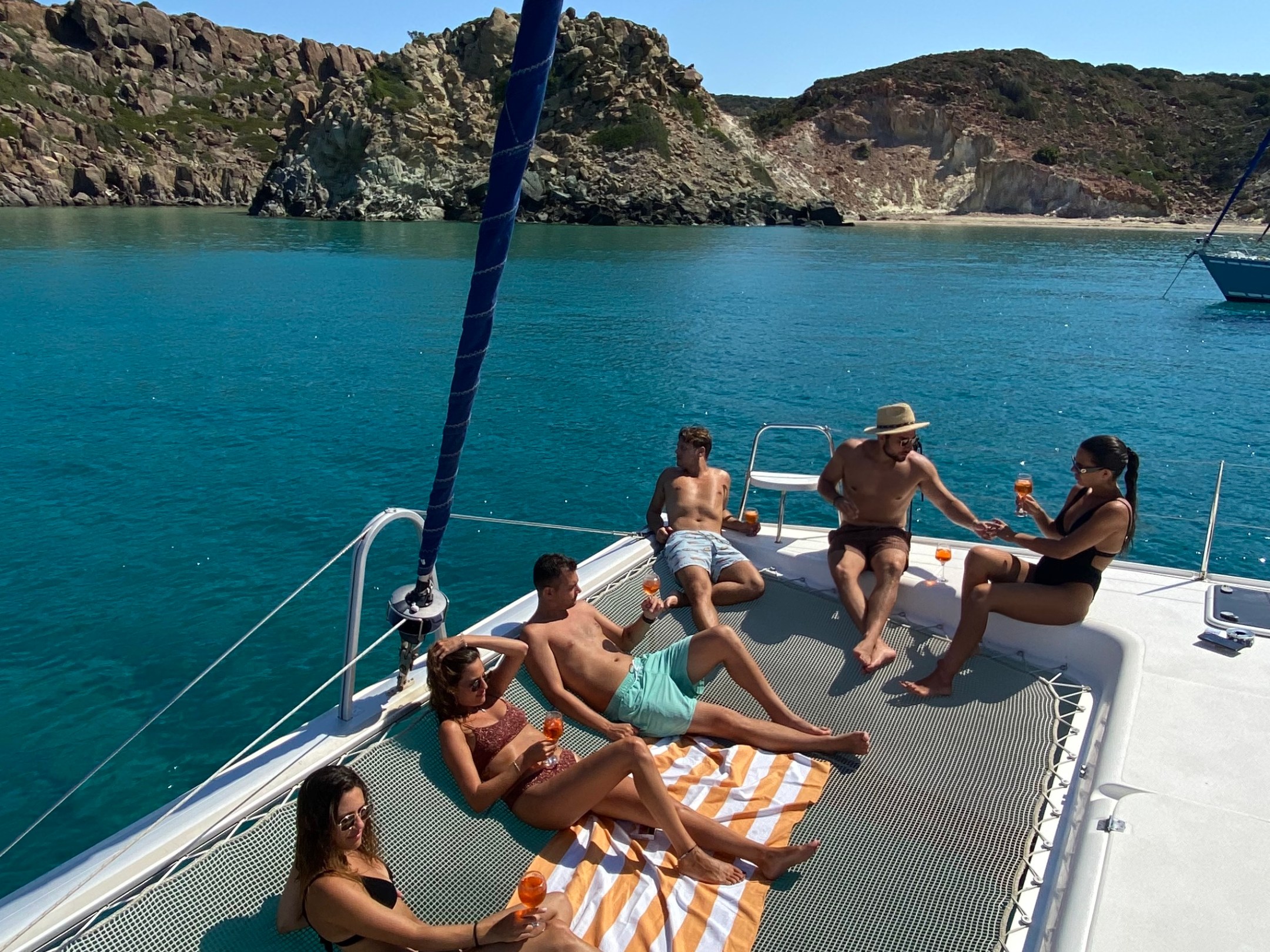 People relaxing on a boat's net hammock with drinks, ocean and rocky cliffs in the background.