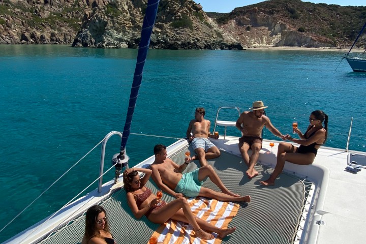 People relaxing on a boat's net hammock with drinks, ocean and rocky cliffs in the background.