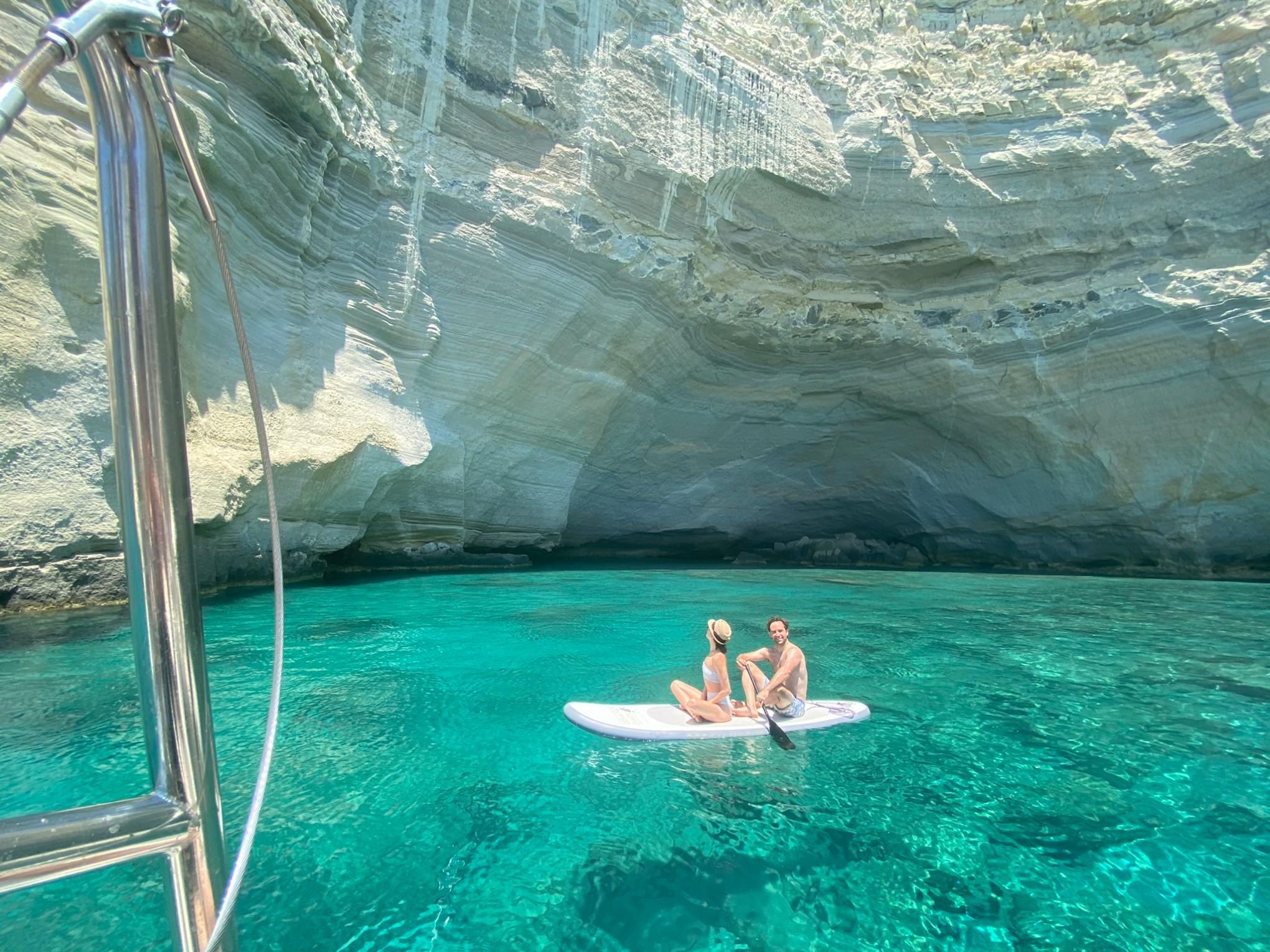 Two people on paddleboard in clear turquoise water near rocky cave entrance.