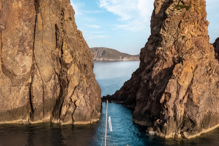 Sailboat passing between two large rock formations in calm blue waters.
