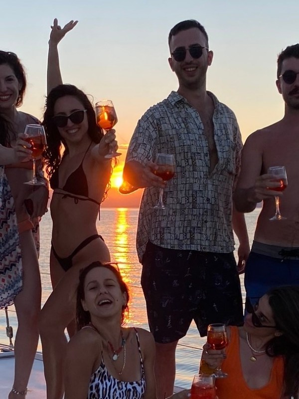 Group of people on a boat at sunset, holding drinks, smiling and posing.