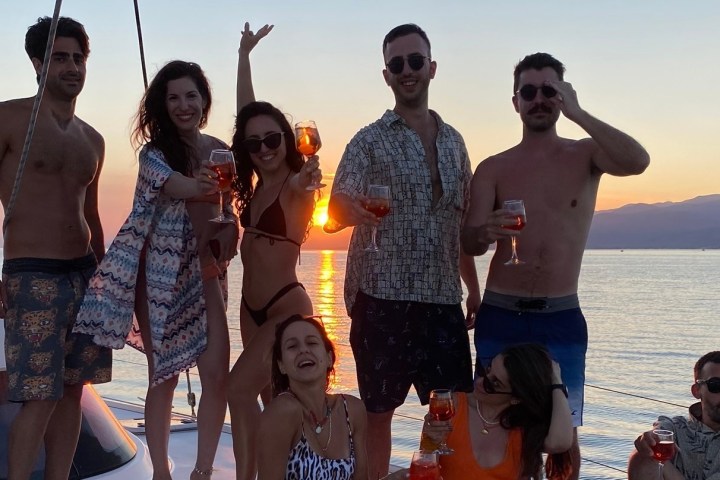 Group of people on a boat at sunset, holding drinks, smiling and posing.