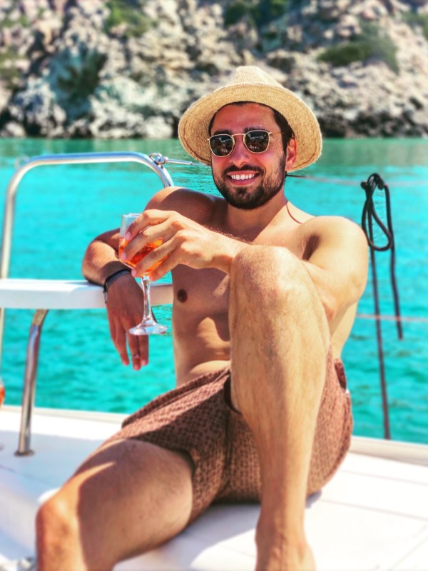 Man in sunglasses and hat relaxes on a boat with a drink, turquoise water and rocky background visible.