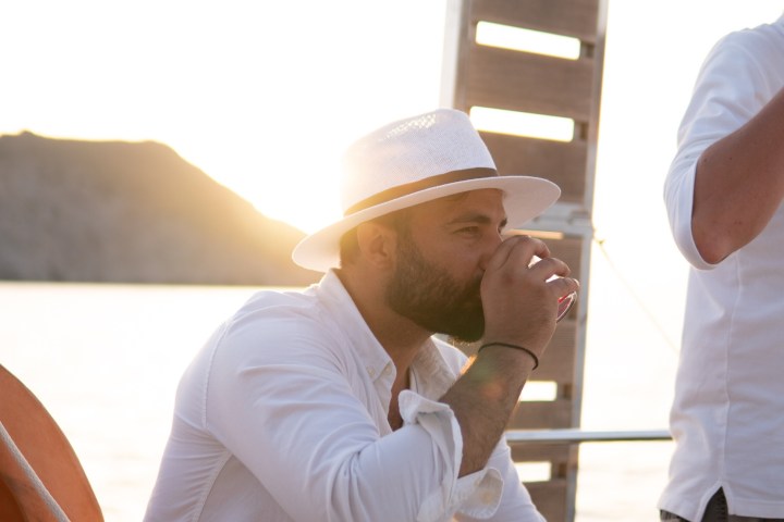 Man in white hat and shirt drinks on a boat at sunset.