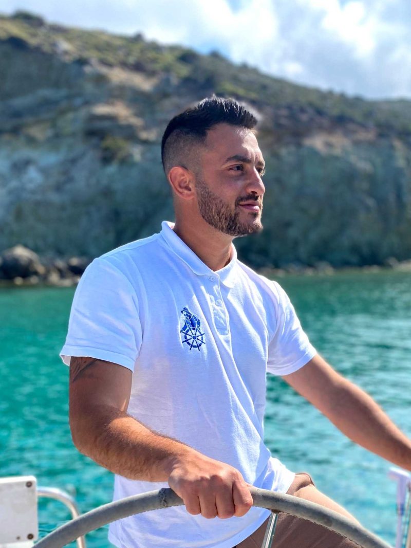 Man in white polo steering a boat with a rocky coastline in the background.