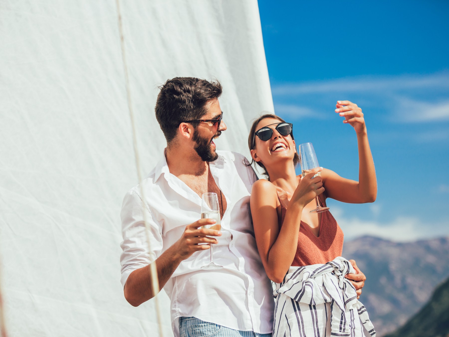 Couple on a sailboat, laughing and holding champagne glasses against a blue sky.