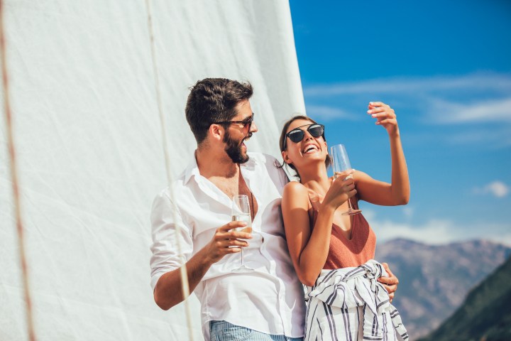 Couple on a sailboat, laughing and holding champagne glasses against a blue sky.