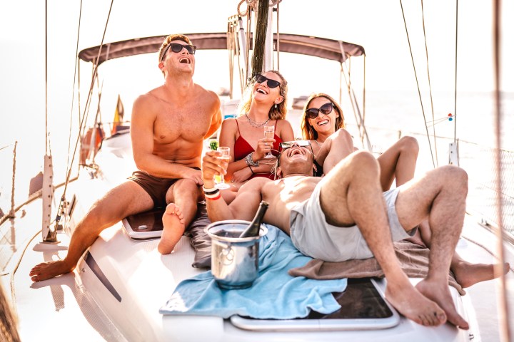 Four friends relaxing and laughing on a boat, holding drinks and enjoying the sun.