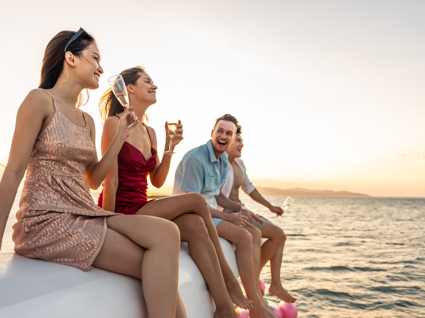 Four people laughing on a boat at sunset, holding drinks, with the ocean in the background.