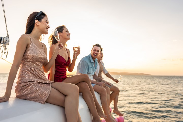 Four people laughing on a boat at sunset, holding drinks, with the ocean in the background.