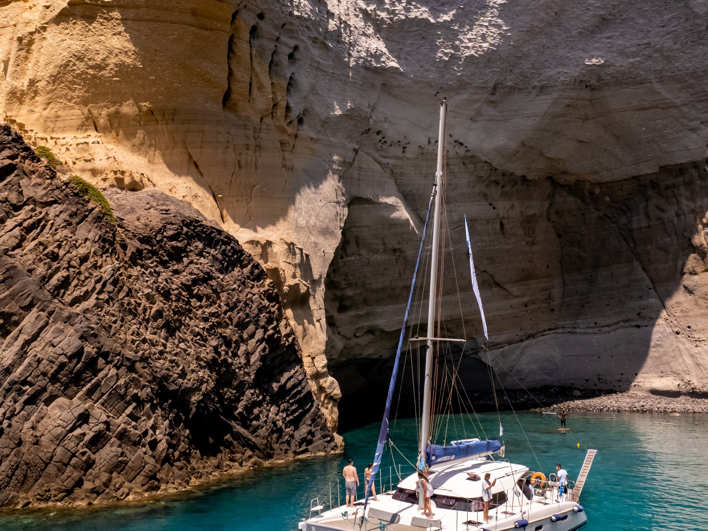 Sailboat in a turquoise cove by tall, textured cliffs under a clear blue sky.