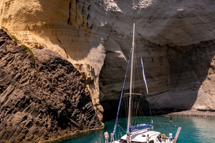 Sailboat in a turquoise cove by tall, textured cliffs under a clear blue sky.