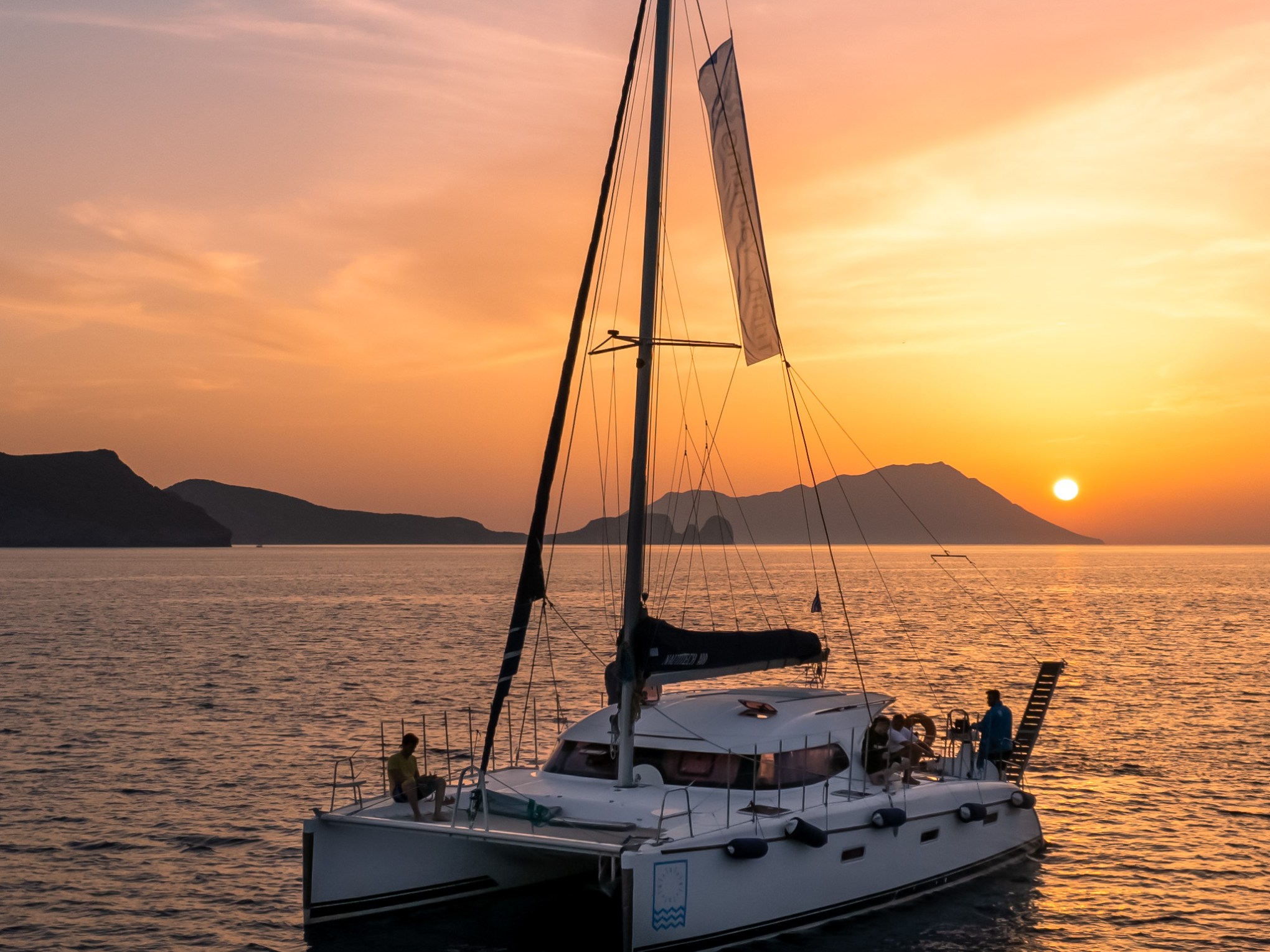 Sailboat on calm sea at sunset, with mountains in the background and colorful sky.