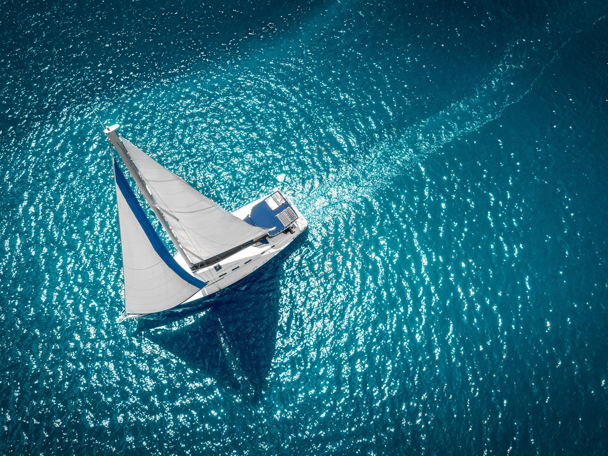 Aerial view of a sailboat on clear blue water with sunlight reflections.